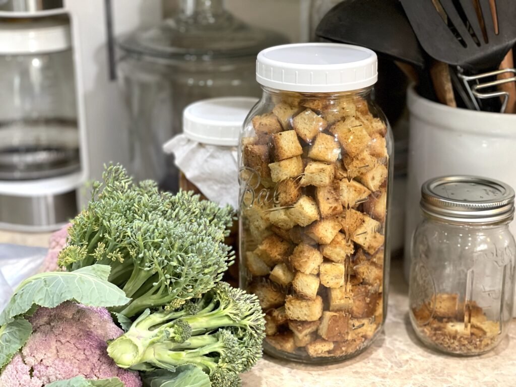 half gallon mason jar filled with homemade croutons on a kitchen counter with fresh broccoli and cauliflower sitting beside it