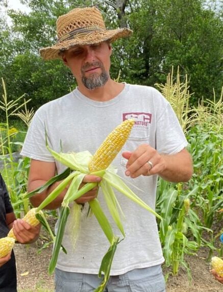 man in front of a corn batch holding freshly picked ears of corn