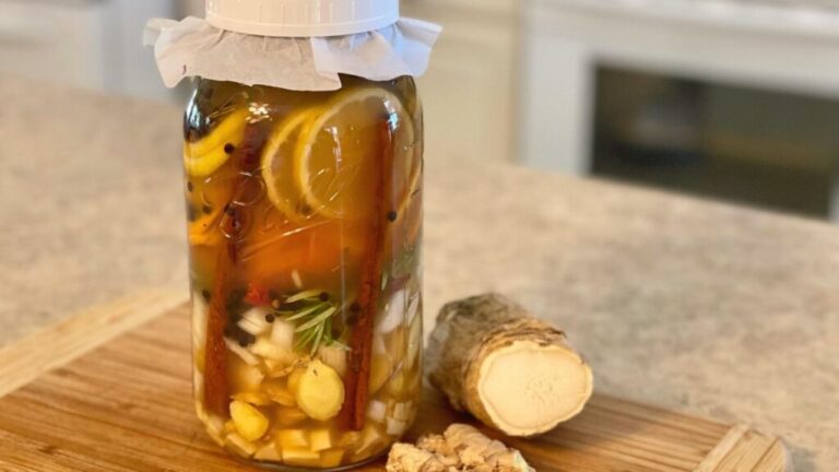 large mason jar filled with fresh ingredients for how to make fire cider with fresh cut ginger laying beside it