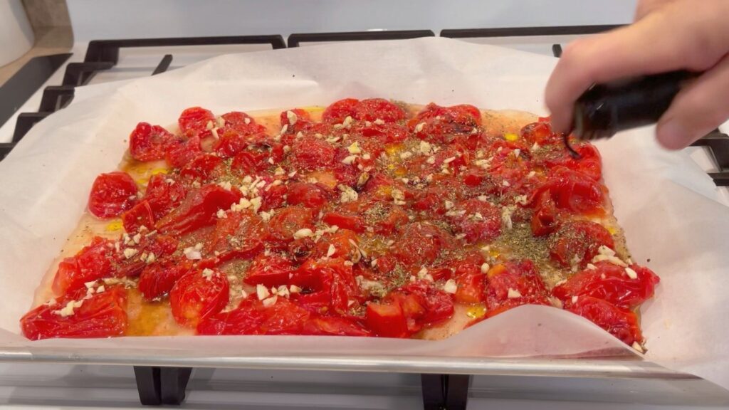 olive oil being drizzled over cut up tomatoes on a baking sheet that has been seasoned with spices and chopped garlic