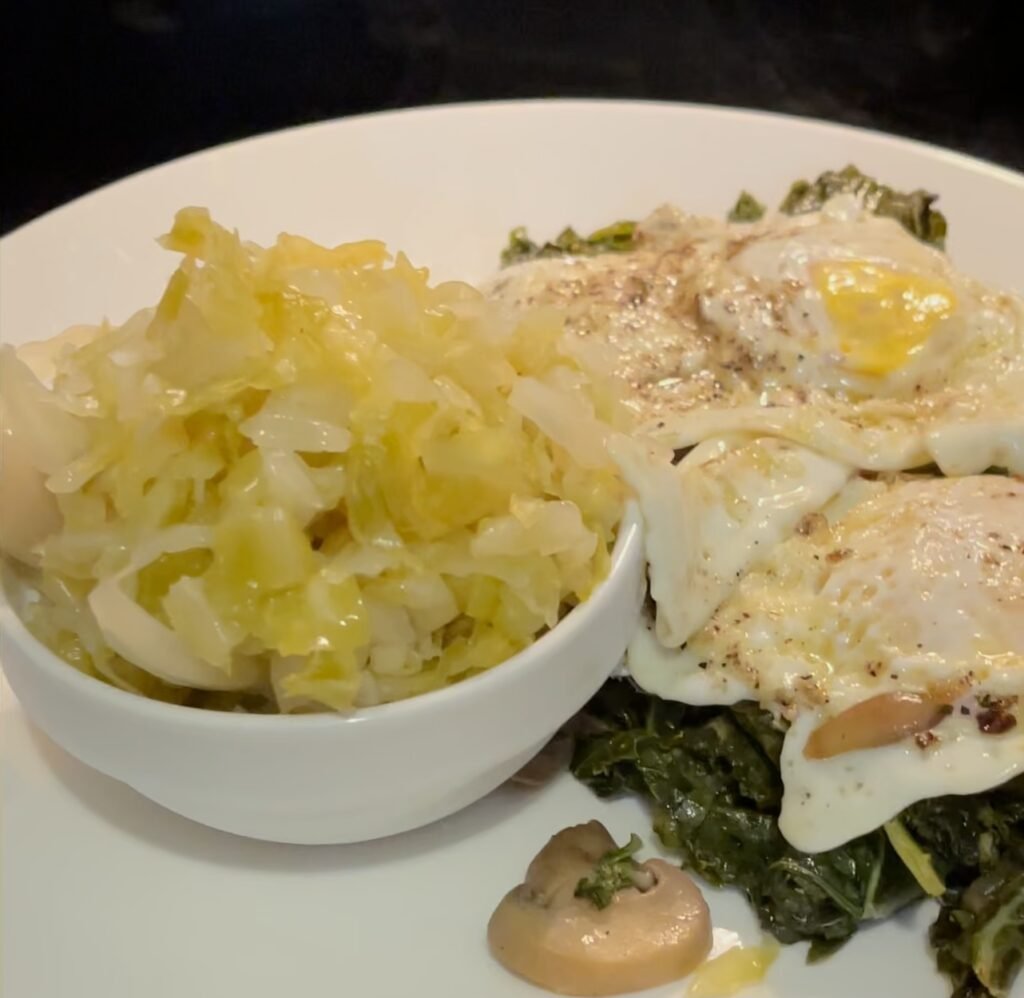 homemade sauerkraut in a small bowl sitting on a plate with sautéed kale and mushrooms beside it with two fried eggs on top